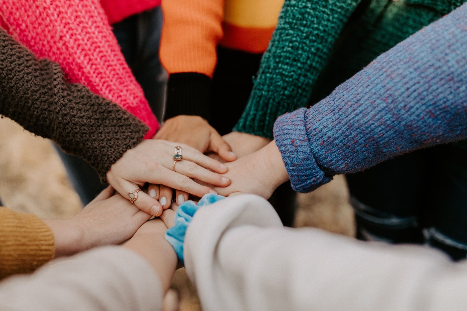 Várias pessoas em um círculo juntam suas mãos. Vemos apenas os braços, com blusas de diferentes cores, e as mãos no centro, formando um símbolo de união.