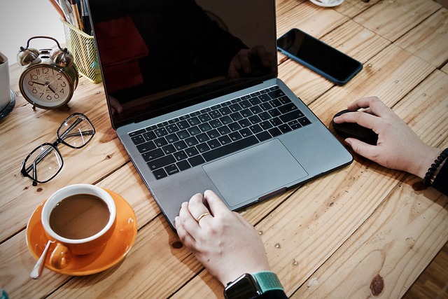 A imagem mostra uma pessoa sentada em uma mesa de jantar de madeira, trabalhando em um laptop. O laptop está posicionado no centro da mesa, e a pessoa está usando um mouse para navegar no dispositivo. Há também dois celulares na mesa, um mais próximo do laptop e outro em direção à borda da mesa.

Além dos dispositivos eletrônicos, há duas xícaras na mesa, uma perto da borda esquerda e a outra perto da borda direita. Um par de tesouras também pode ser visto na mesa, mais próximo ao lado esquerdo. A pessoa parece estar focada em seu trabalho, cercada por vários itens na mesa.