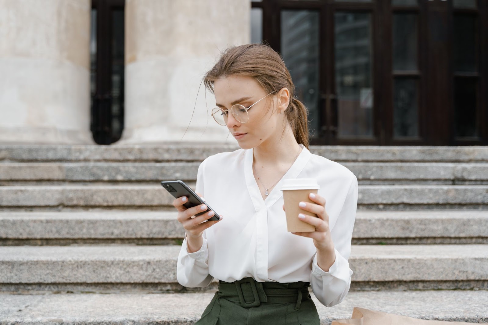 A imagem mostra uma mulher em frente a uma escada, segurando um celular em uma mão e uma xícara de café na outra. Ela parece estar concentrada em seu celular, possivelmente verificando mensagens ou navegando na internet. A mulher está vestindo uma camisa branca e uma saia verde, o que adiciona um toque de cor à cena. A xícara de café está colocada na frente dela, e ela parece estar desfrutando de sua bebida enquanto usa o celular.