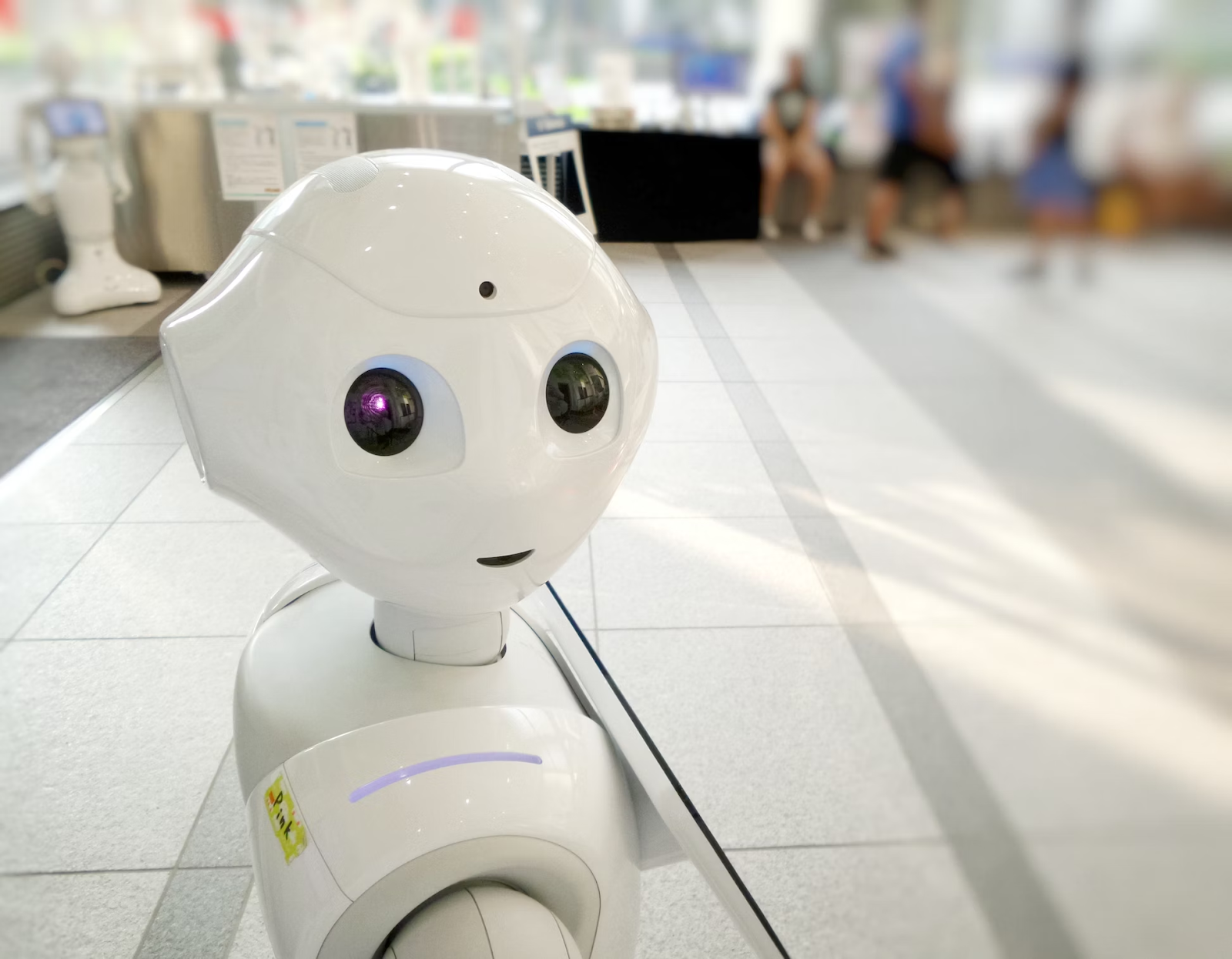 Robô com formas humanóides brancoe sorridente. No fundo há outro robô, pessoas andando e sentadas em uma vitrine de loja