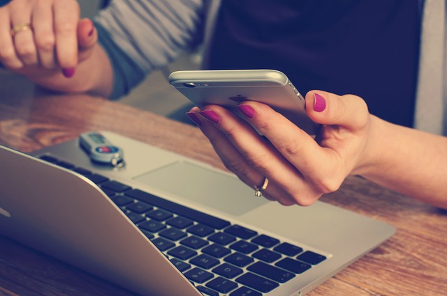 A imagem mostra uma pessoa sentada à mesa com um laptop e um celular. A pessoa segura o celular na mão, possivelmente enviando mensagens ou navegando na internet. Há também uma chave de carro na mesa, sugerindo que a pessoa pode ter acabado de chegar ao seu destino ou está se preparando para sair. A mão da pessoa também é visível na cena, segurando o celular.