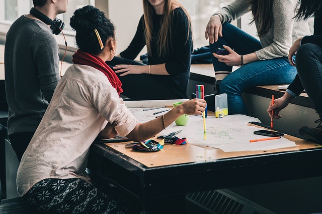 A imagem mostra um grupo de pessoas reunidas ao redor de uma mesa, envolvidas em uma atividade colaborativa. Há um total de seis pessoas, algumas sentadas em um banco e outras em cadeiras. Elas estão trabalhando em um projeto juntas, com vários itens na mesa, como uma xícara, um celular, um lápis e um par de tesouras.

A mesa está cheia de papéis, e há dois livros colocados sobre a mesa também. As pessoas do grupo estão focadas em sua tarefa, e a atmosfera parece ser cooperativa e produtiva.