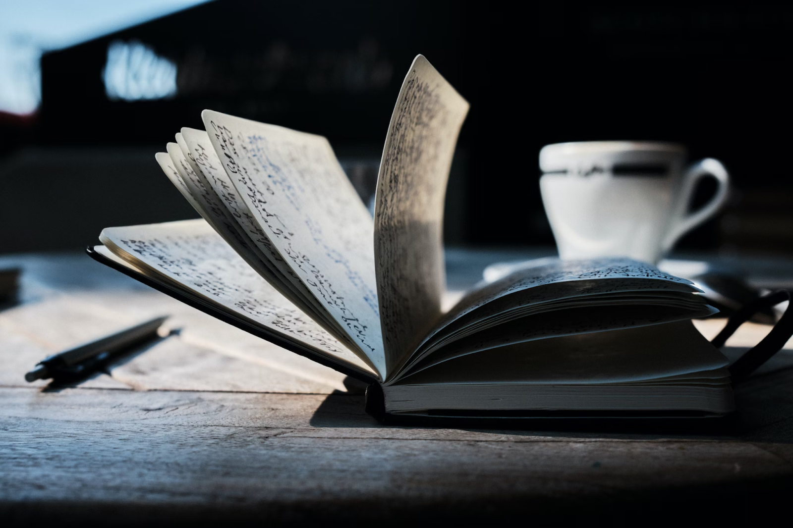 Mesa de madeira com um caderno aberto e várias páginas escritas à mão. A mesa também possui uma xícara posicionada perto do canto superior direito.