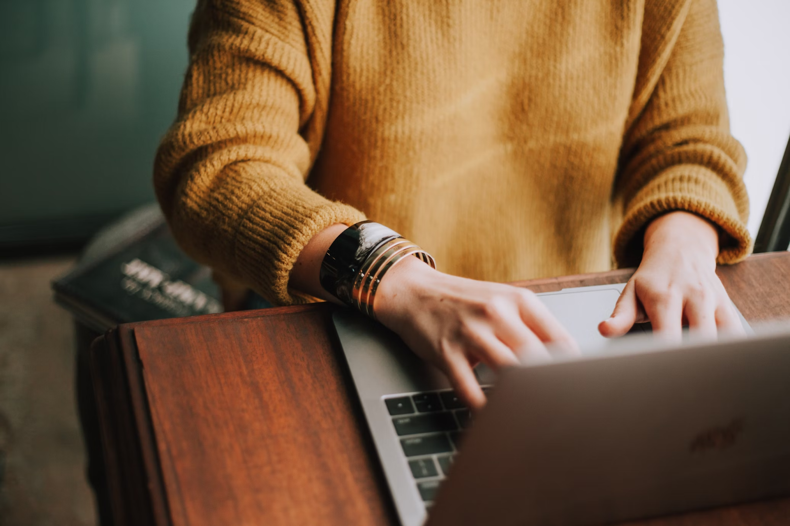 Pessoa sentada em uma mesa de madeira, utilizando um computador portátil. A pessoa está vestindo um suéter amarelo, e suas mãos estão visíveis no teclado do laptop.