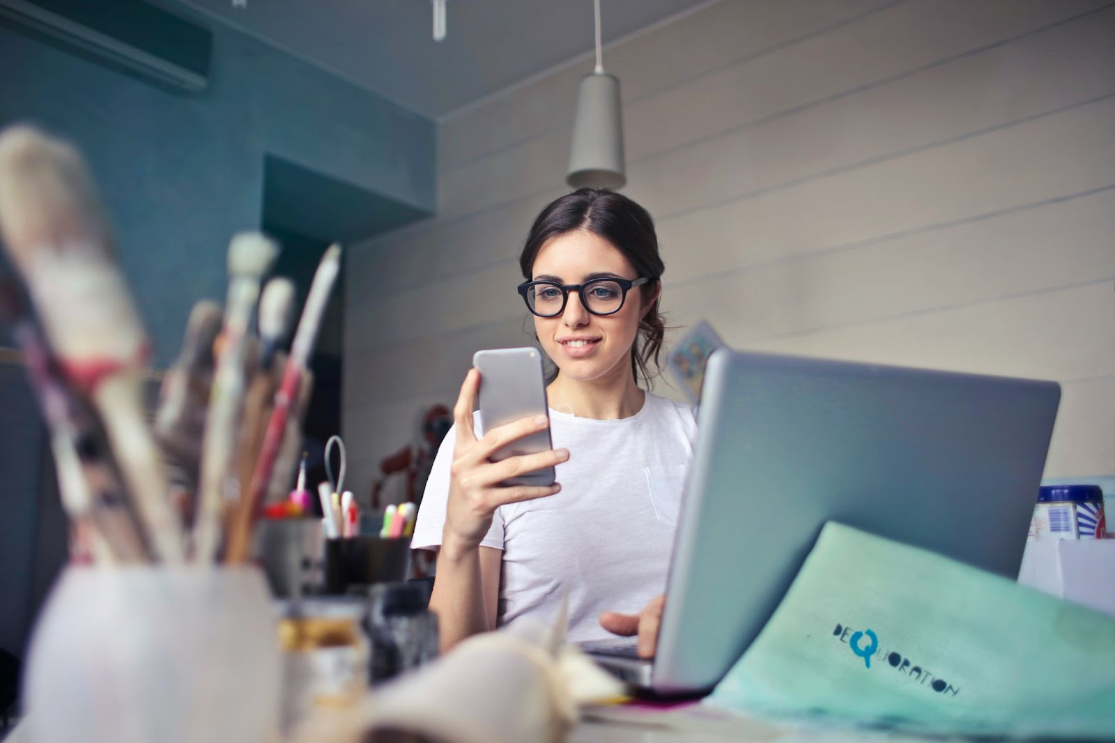 A imagem retrata uma mulher sentada em uma mesa, segurando um celular em sua mão. Ela também está usando um laptop, que está posicionado na mesa à sua frente. A mulher parece estar concentrada em seu trabalho ou interagindo com o celular.

A mesa está bem equipada com vários itens, incluindo uma xícara, uma tigela e uma colher. Também há vários livros espalhados pela mesa, possivelmente para referência ou leitura recreativa. Uma cadeira está posicionada em frente à mesa, proporcionando uma área de assento confortável para a mulher.