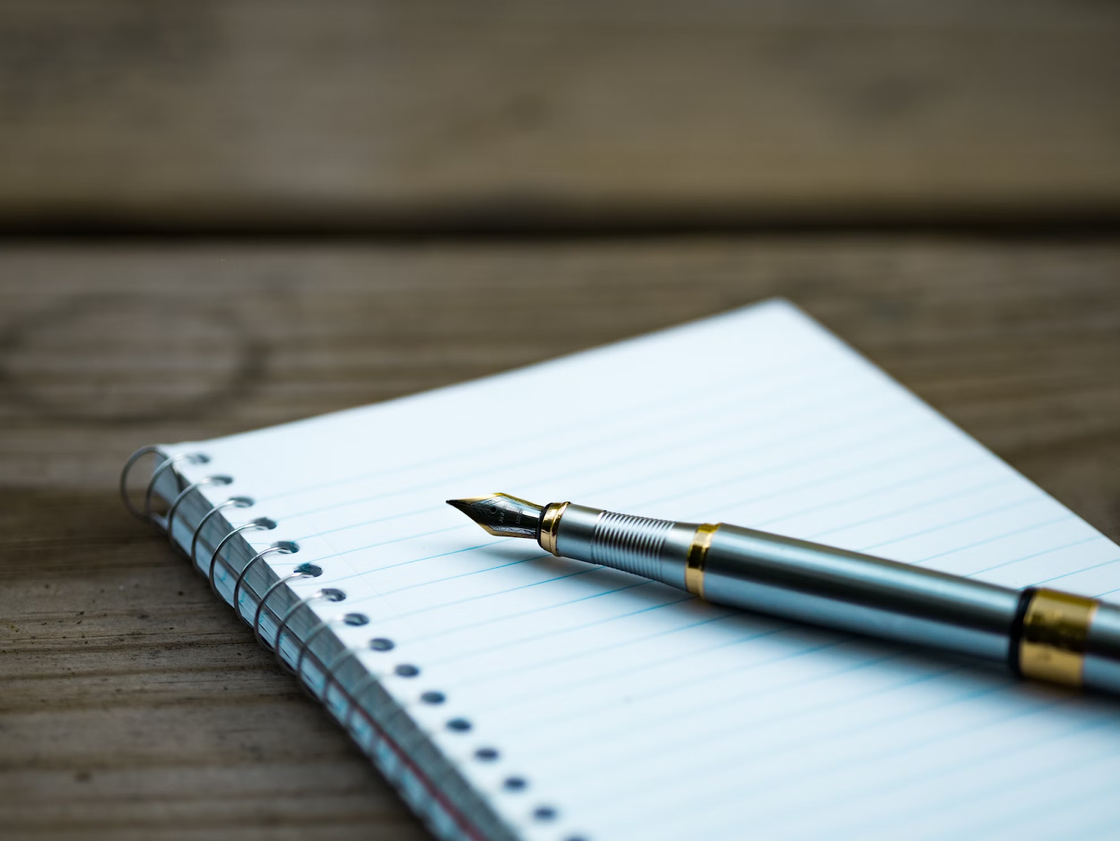 Caneta sobre um caderno com pautas e folhas brancas. Estes itens estão em cima de uma mesa.