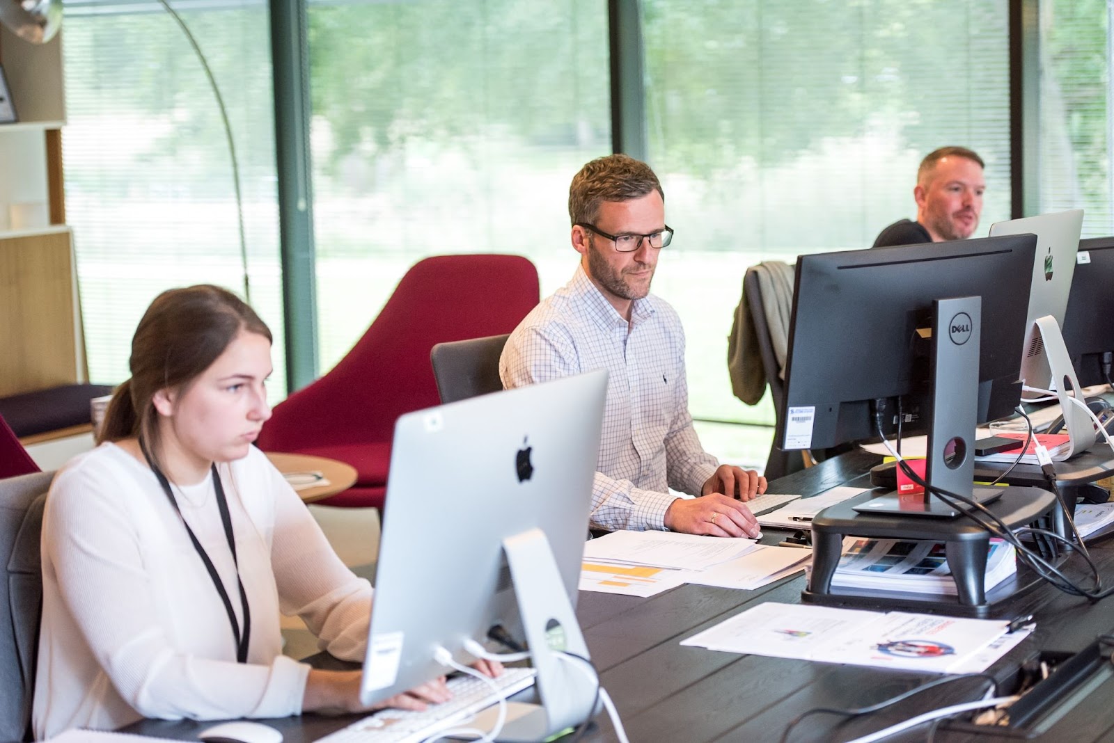 A imagem retrata um grupo de três pessoas sentadas em uma mesa longa, trabalhando em seus computadores. Duas das pessoas estão focadas em suas tarefas, enquanto a terceira pessoa parece estar observando algo diferente. Cada pessoa tem um computador na frente delas, sendo que uma pessoa está usando um laptop e as outras duas estão usando computadores de mesa.

A mesa está cercada por cadeiras, com uma cadeira do lado esquerdo, outra do lado direito e uma terceira cadeira no final da mesa. Também são visíveis dois teclados na mesa, um próximo ao laptop e outro mais próximo do computador de mesa do lado direito. Um livro também pode ser visto na mesa, possivelmente para referência ou leitura de lazer.