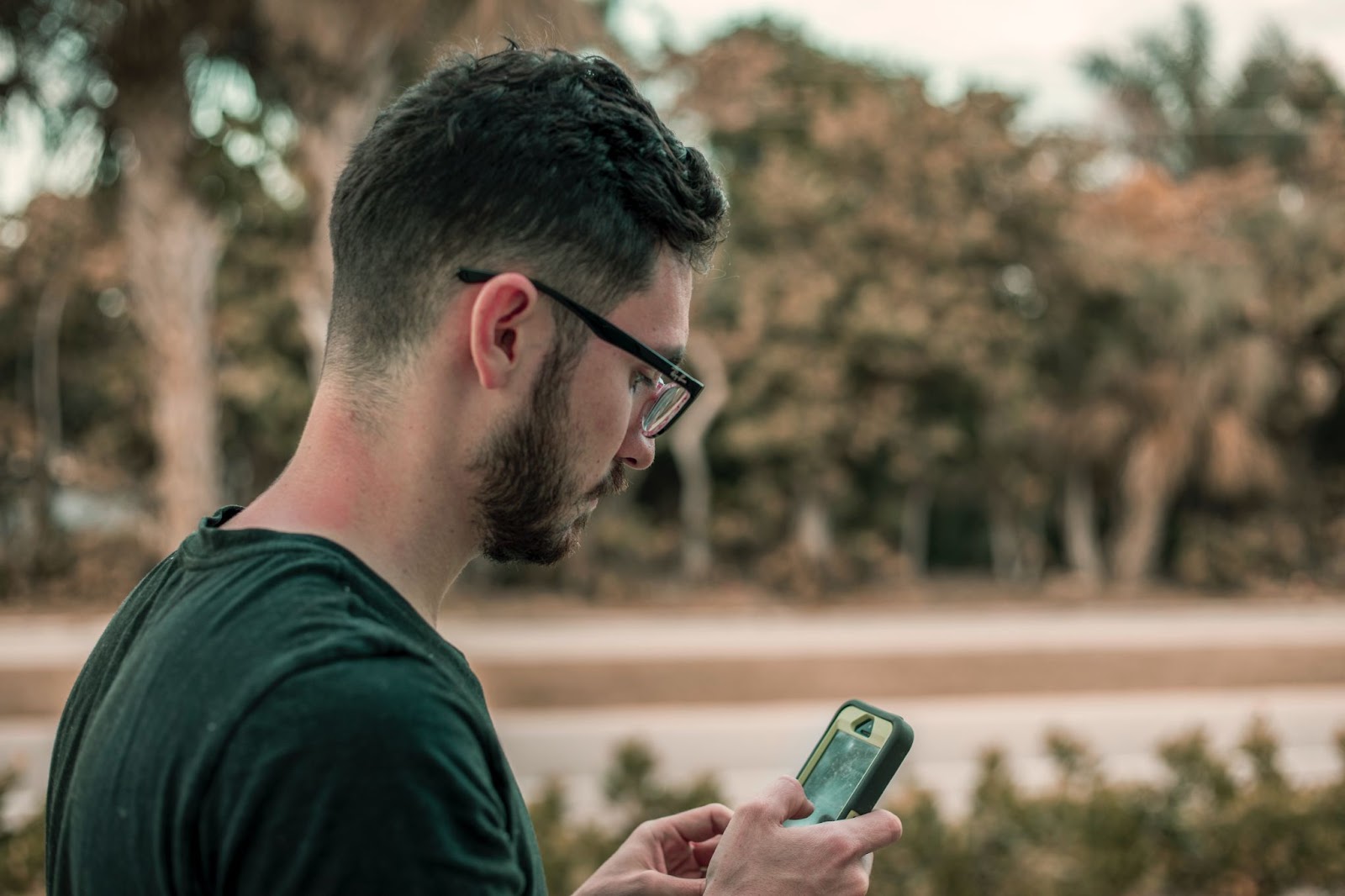 A imagem mostra um homem usando óculos e uma camisa preta, parado do lado de fora e olhando para o seu celular. Ele parece estar focado na tela, possivelmente checando mensagens ou usando algum aplicativo. O homem está posicionado próximo ao centro da cena, com o celular em suas mãos.

Ao fundo, é possível ver várias árvores, fornecendo um ambiente natural para a atividade ao ar livre do homem. A roupa que ele veste e a presença do celular sugerem que ele pode estar fazendo uma pausa no trabalho ou desfrutando de algum tempo livre ao ar livre.