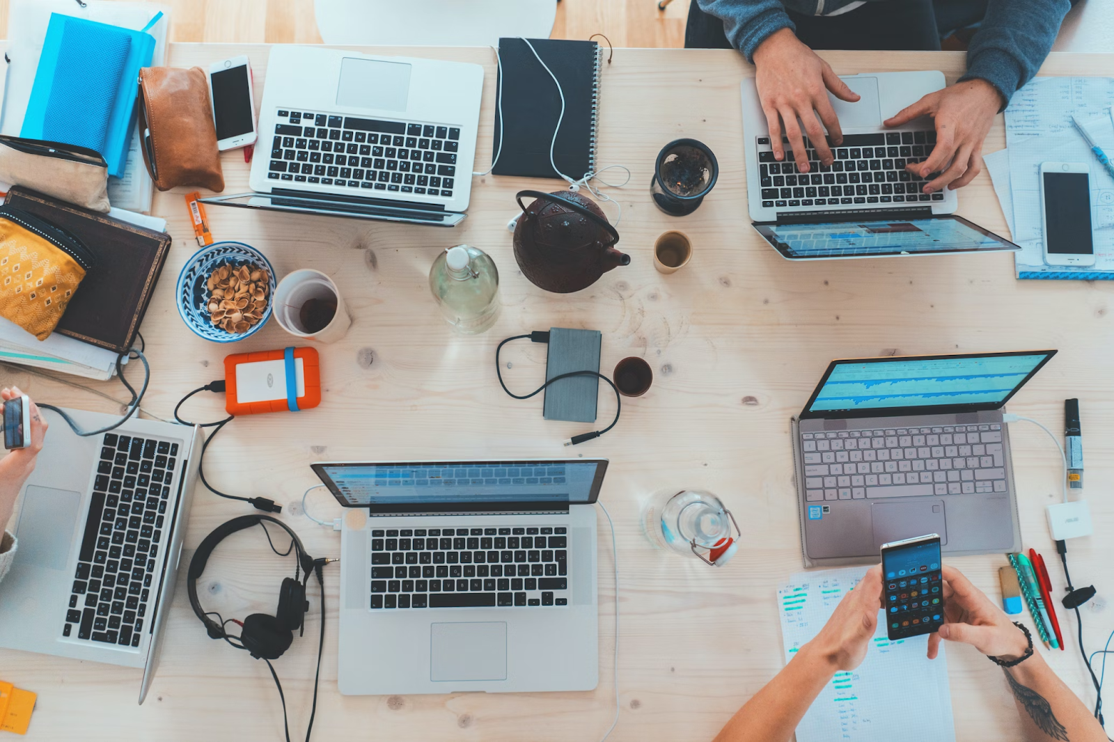 pessoas interagindo com aparelhos eletrônicos em meio a uma mesa lotada por notebooks, itens tecnológicos, itens pessoais e itens de escritório.