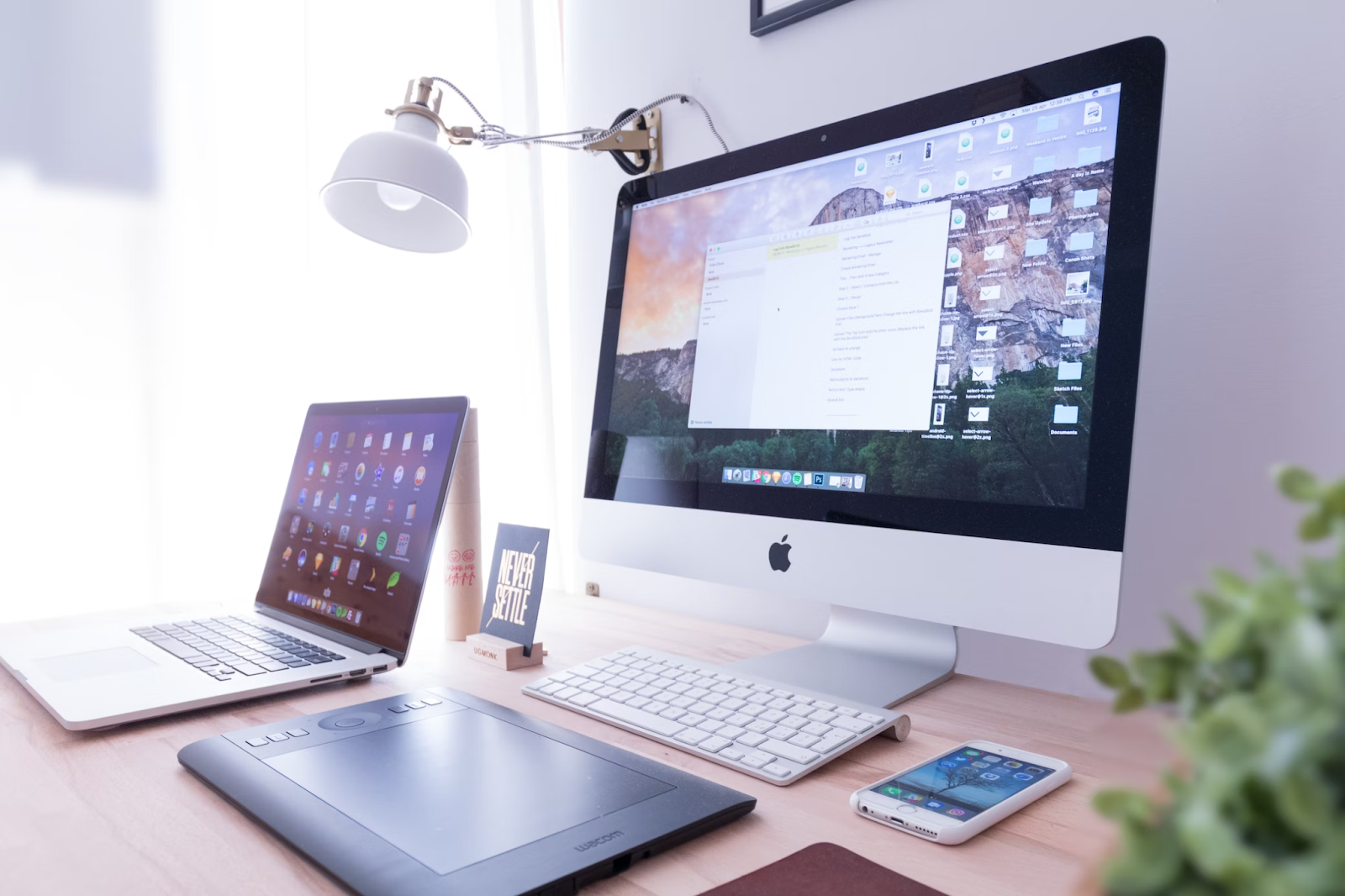 na imagem, uma mesa de escritório com notebook, mesa digitalizadora, iPhone e um iMac. A imagem é bem iluminada.