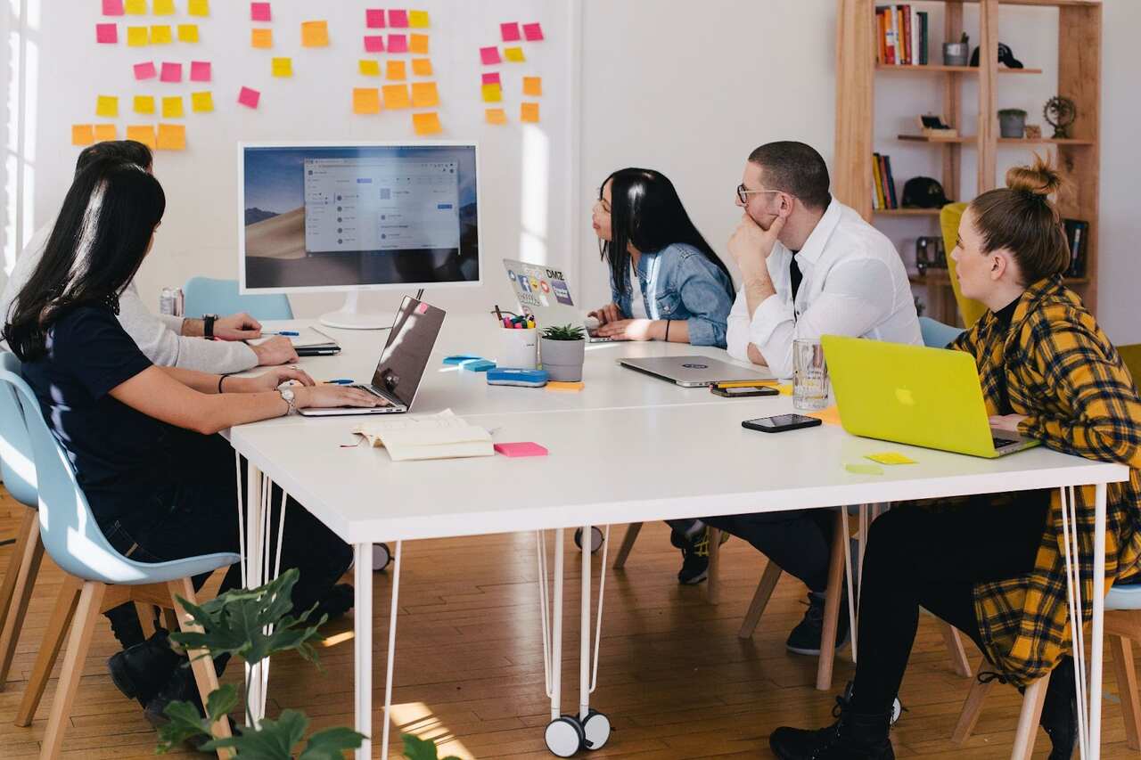 Cinco pessoas sentadas ao redor de uma mesa olhando para uma tela. Cada pessoa tem seu próprio computador, e a mesa contém celulares, cadernos, canetas e post-its. A parede branca ao fundo também está coberta de post-its.