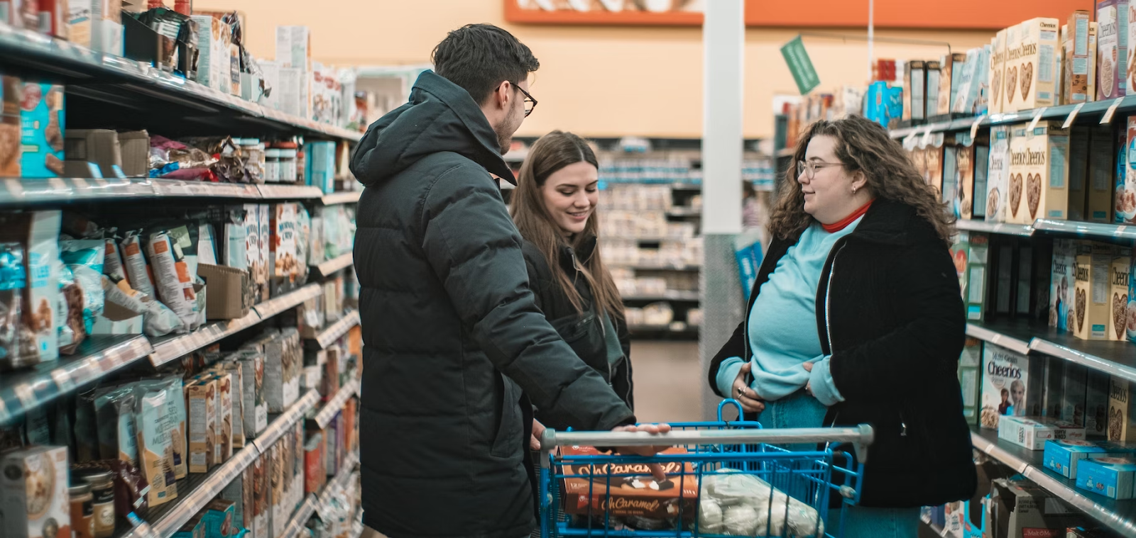 três pessoas ao redor de um carrinho de compras em um corredor de produtos de um supermercado.