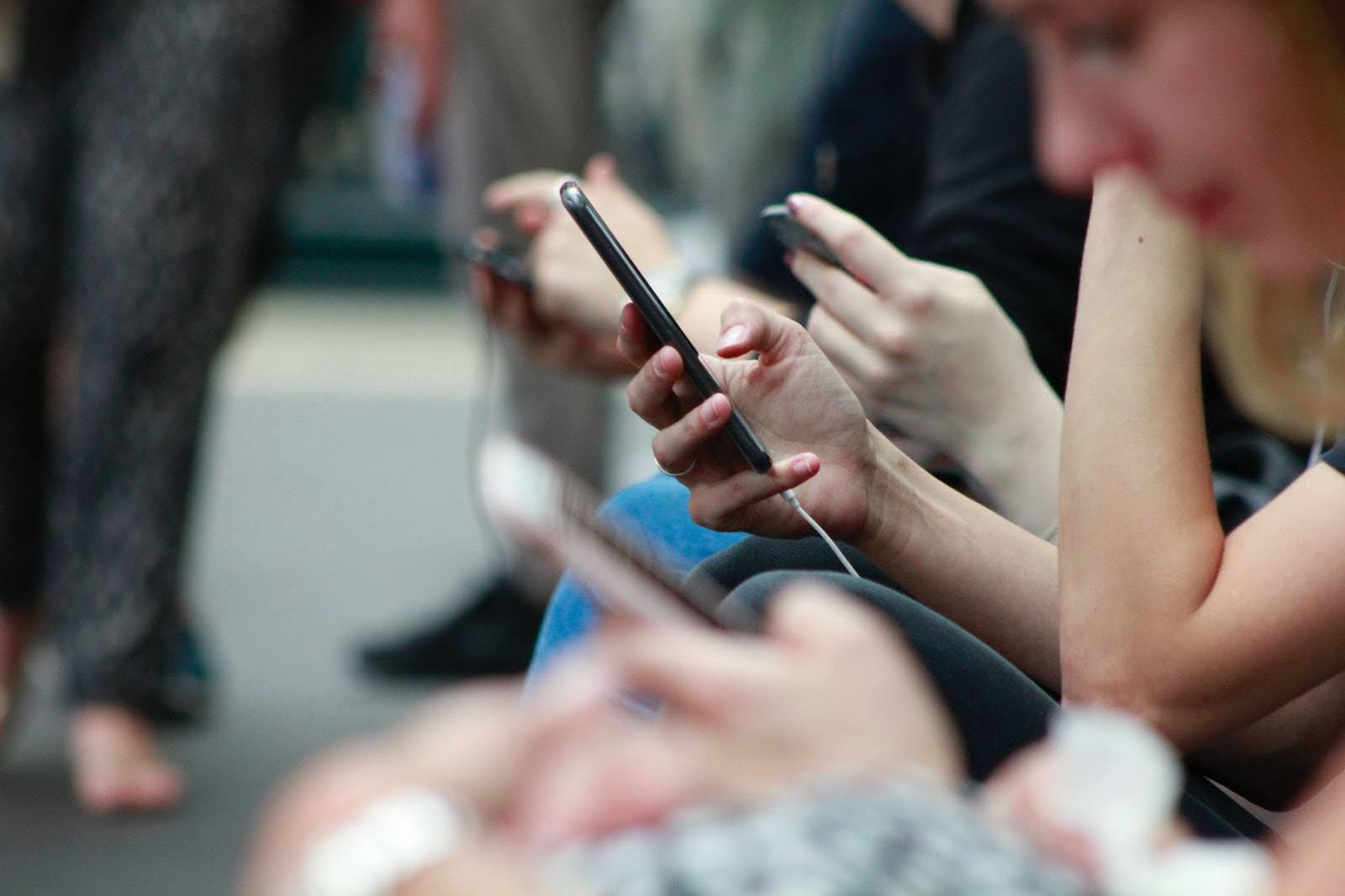 A imagem mostra um grupo de pessoas sentadas uma ao lado da outra, todas elas absortas em seus celulares. Há pelo menos cinco celulares visíveis na cena, com cada pessoa segurando seu dispositivo em suas mãos. As pessoas estão sentadas próximas umas das outras, com algumas sentadas em um banco e outras em uma cadeira.
Além das pessoas e dos celulares, há dois relógios na cena, um localizado perto do canto superior esquerdo e outro no meio da imagem. A presença dos relógios sugere que as pessoas podem estar esperando por algo ou acompanhando o tempo enquanto usam seus celulares.
