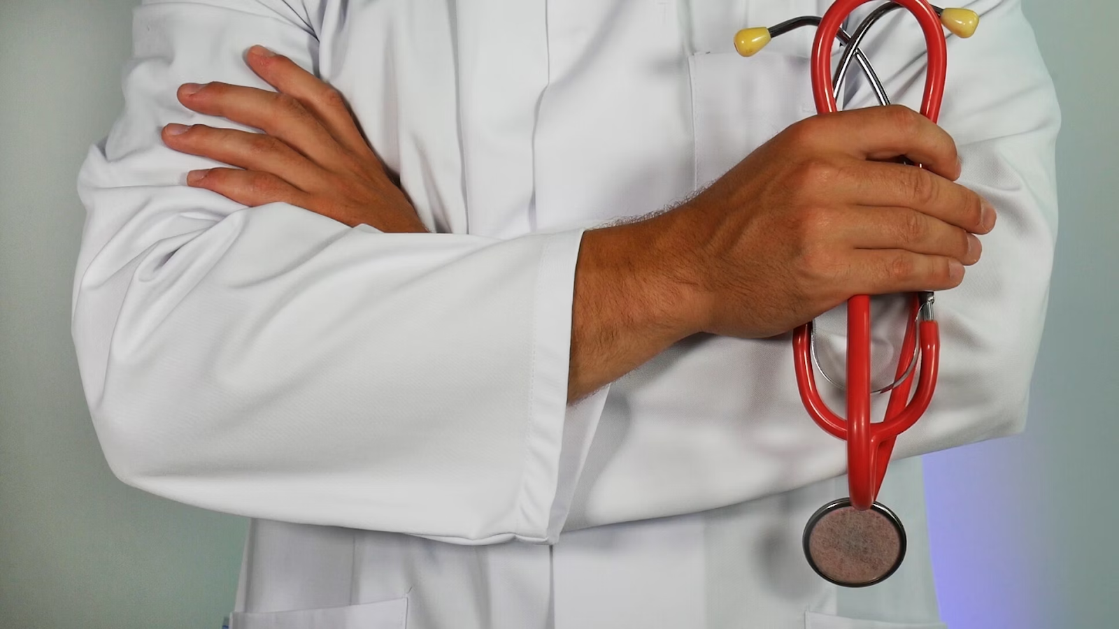 Médico trajando jaleco, cruzando braços e segurando um estetoscópio em uma das mãos.